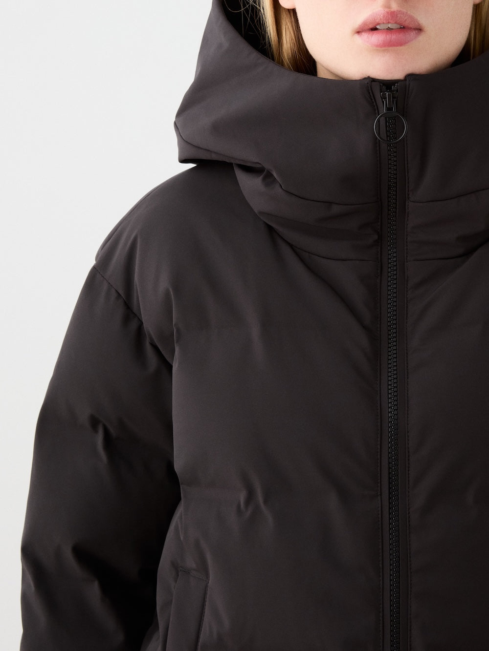 A close-up shows a person in the We Norwegians Aurora Ski Jacket women, a black puffer with responsible down, large hood, and front zipper; only the lower part of their face is visible.