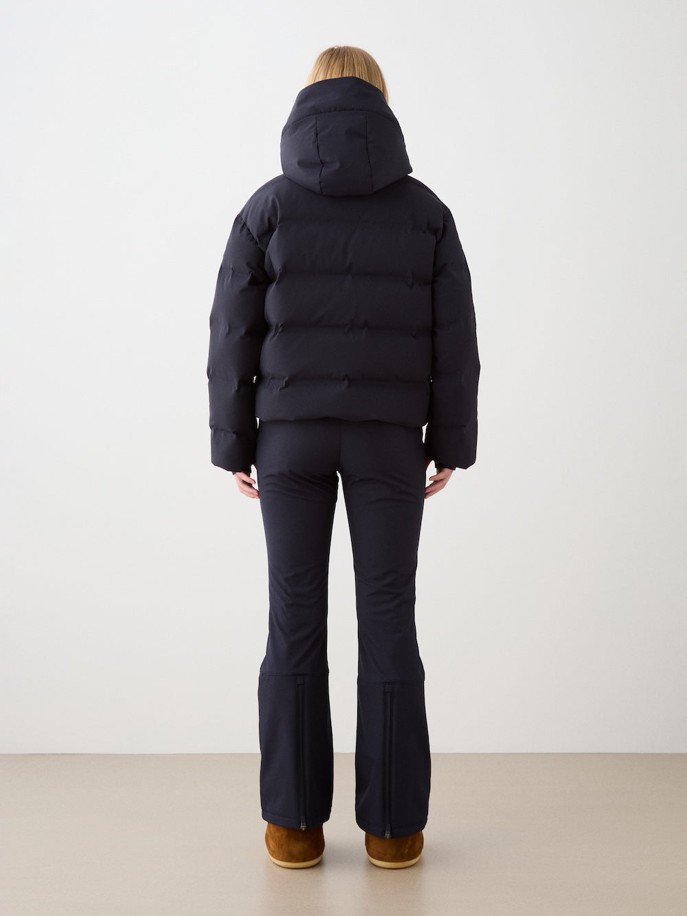 A person stands facing away in a puffy black We Norwegians Aurora Ski Jacket with responsible down, paired with fitted black flared pants and brown shoes, set against a plain light-colored background.