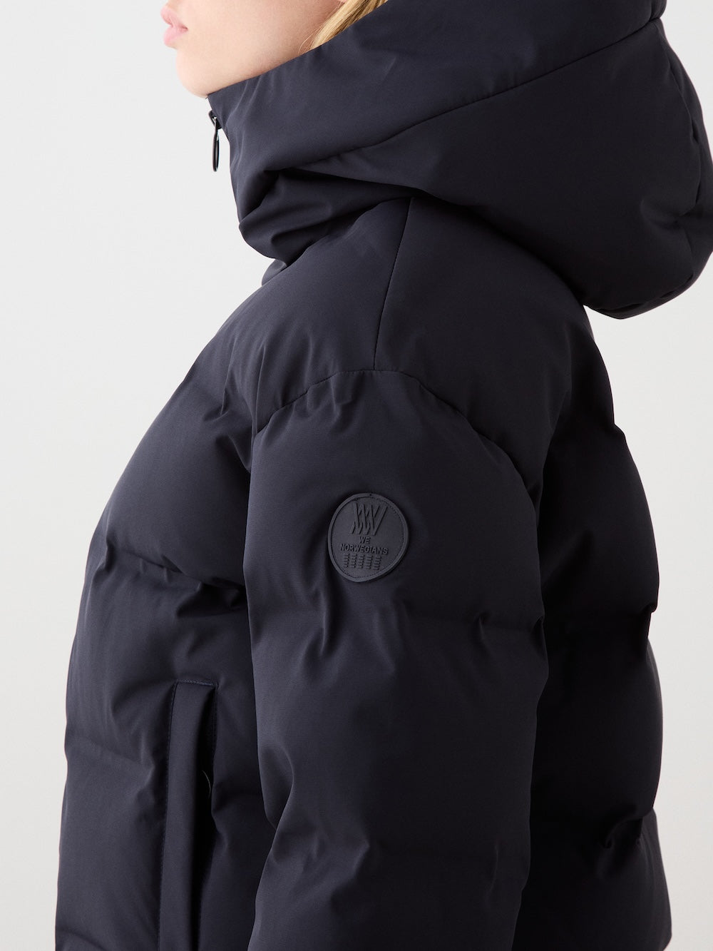 A woman is shown from the side wearing a black We Norwegians Aurora Ski Jacket, featuring a high collar, thick waterproof material, and a patch on the upper sleeve. The background is plain and light-colored.