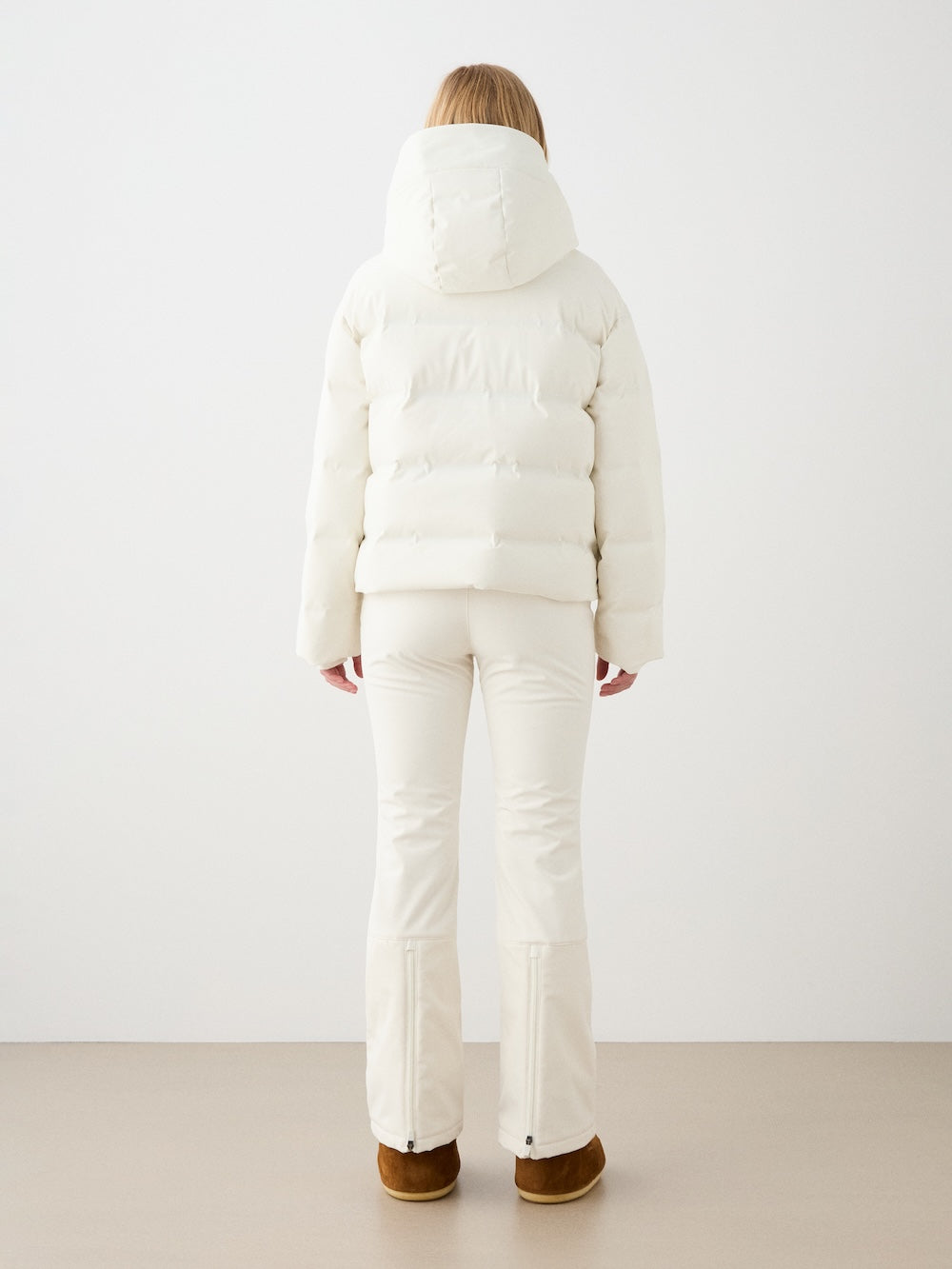 A person with long hair stands facing away, wearing a hooded white We Norwegians Aurora Ski Jacket women with responsible down, paired with white pants and brown boots against a plain light background.
