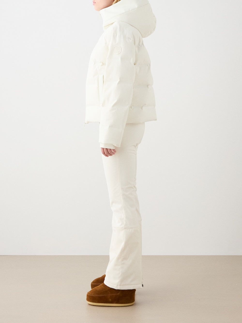 A person stands sideways in a We Norwegians Aurora Ski Jacket for women—puffy and white, made with responsible down—paired with white pants and brown winter boots. Their face isn't visible; the background is plain.