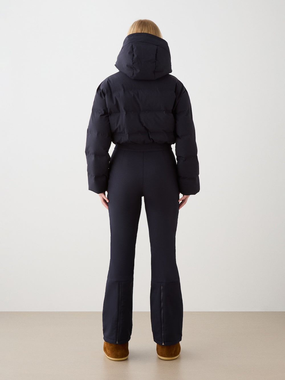A woman with blonde hair, seen from behind, wears the Aurora Ski Suit by We Norwegians with brown boots, standing against a plain light background.