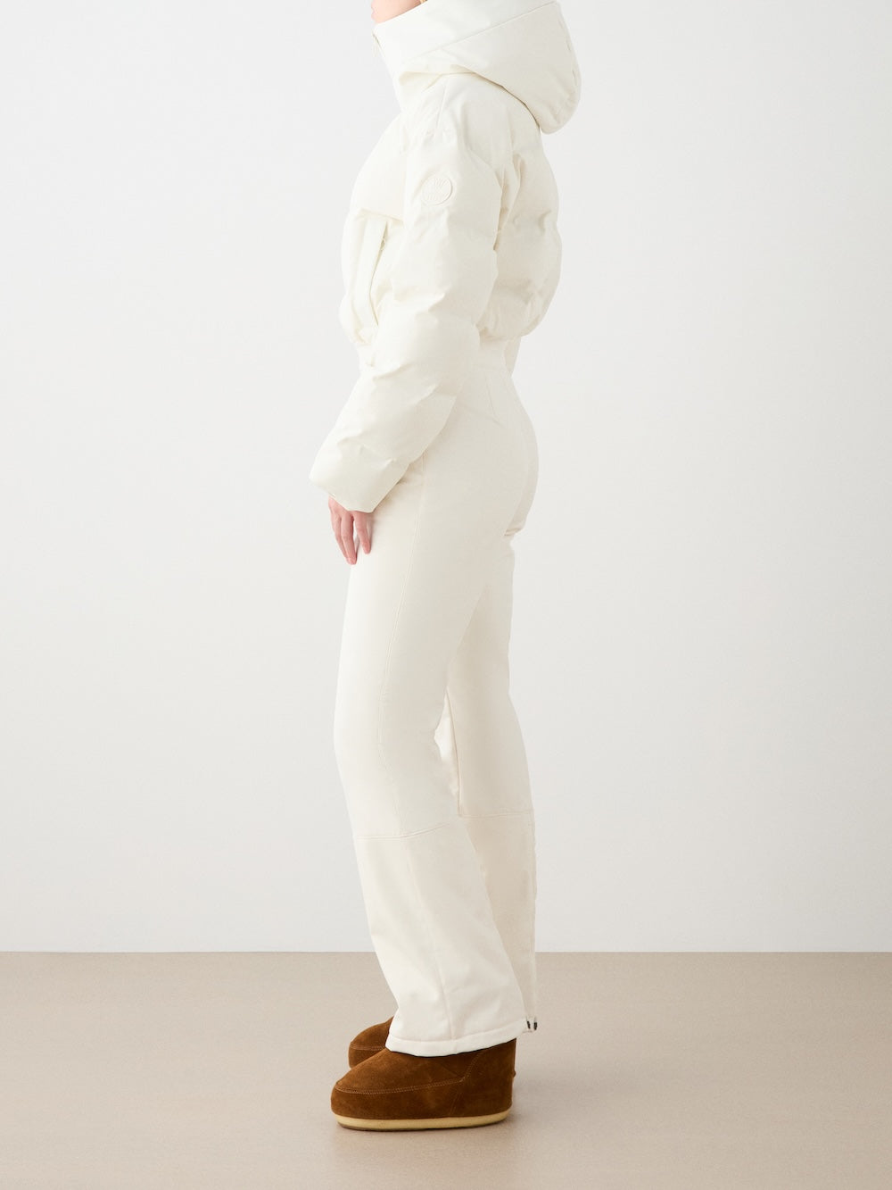 A person in profile models We Norwegians’ fitted Aurora Ski Suit women in all-white with a hood and responsible down, paired with brown winter boots, set against a plain light background for a refined alpine look.