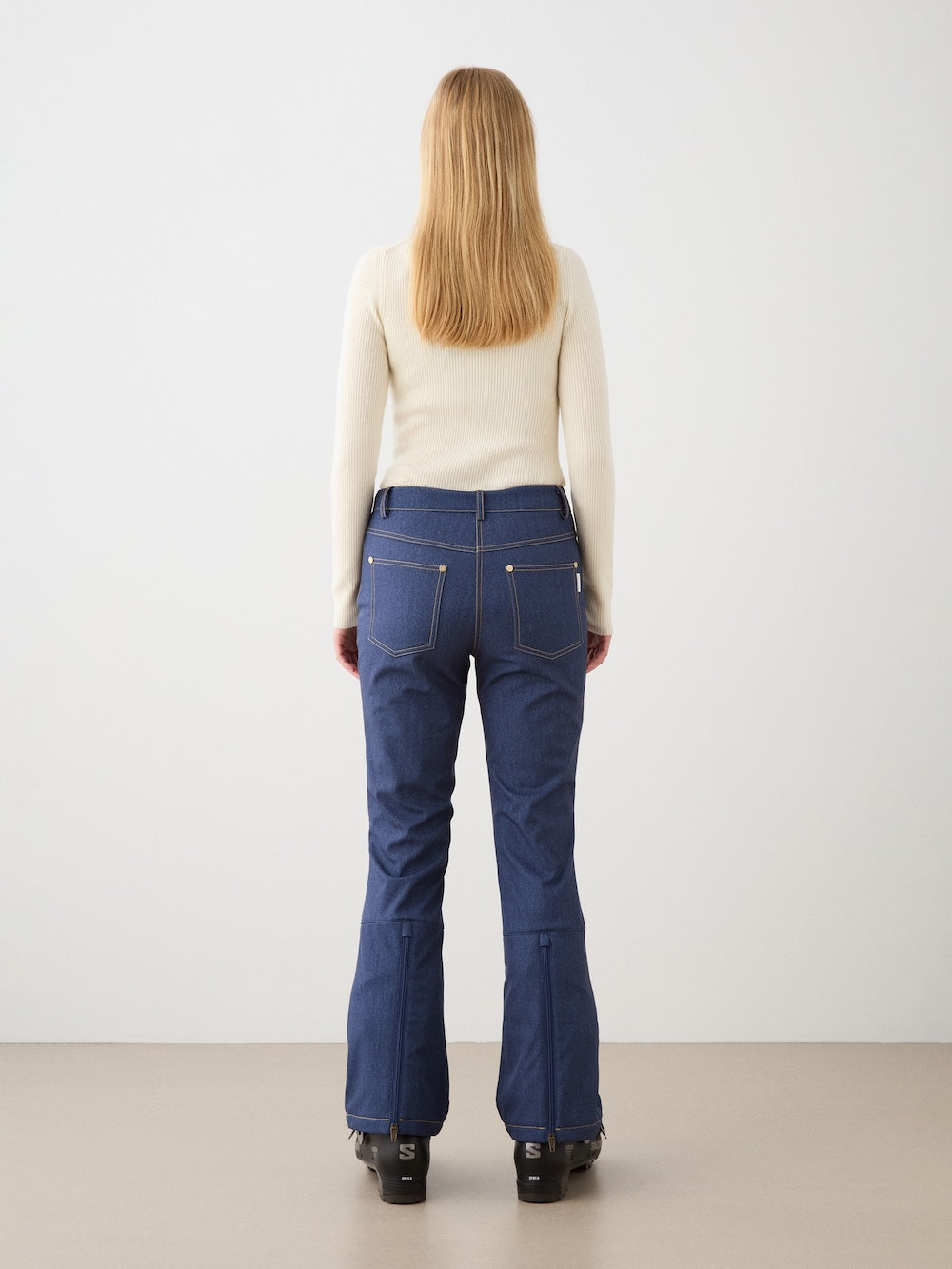 A woman with straight blonde hair, facing away from the camera, wears a cream long-sleeve top and We Norwegians Western Ski Pant in blue denim with visible stitching and black shoes, set against a plain light background.