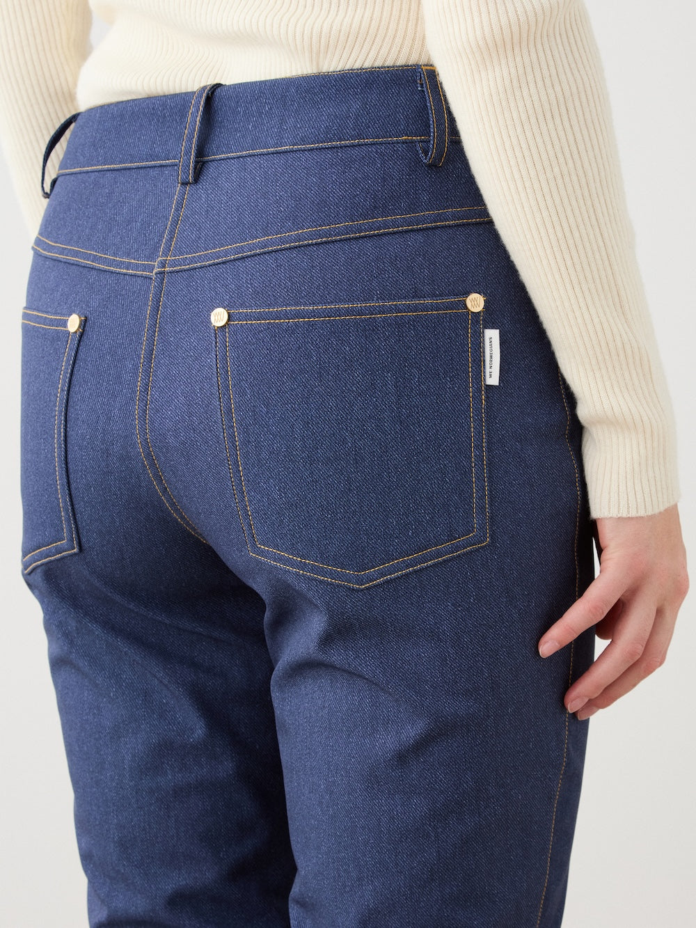 A woman wears We Norwegians’ Western Ski Pant in blue denim with rodeo-inspired stitching, paired with a cream ribbed sweater. Shown from the waist down and back, one hand rests near the right pocket, highlighting fabric texture.