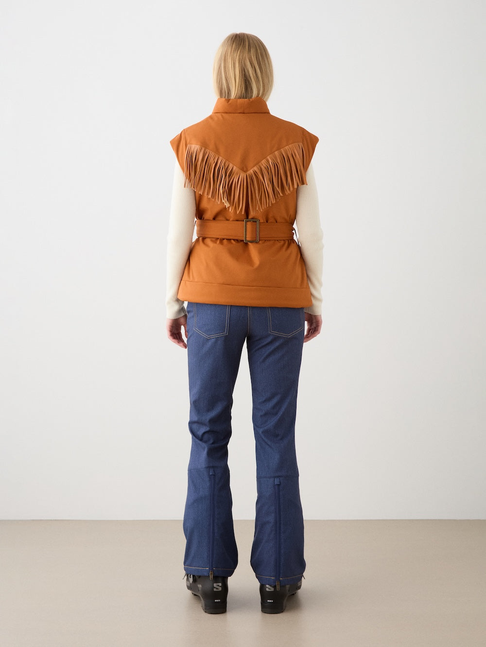 A person stands facing away, wearing the We Norwegians Western Vest women in rust-orange with fringe and a belt, paired with a cream long-sleeve top, blue jeans with visible seams, and black shoes against a plain background.