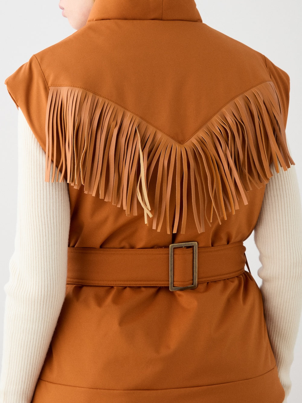A woman wears the We Norwegians Western Vest—a sleeveless, belted, rust-hued vintage ski vest with long fringe across the back—over a cream ribbed long-sleeve top, pictured from shoulders to waist.