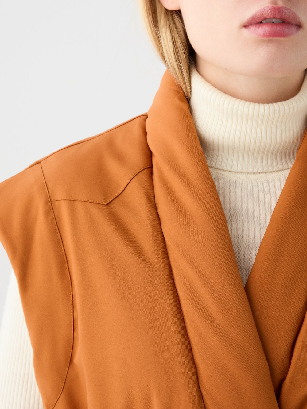 A close-up shows a person wearing a cream turtleneck and the We Norwegians Western Vest women in rust, featuring a wide collar; only the lower half of their face and upper torso are visible.