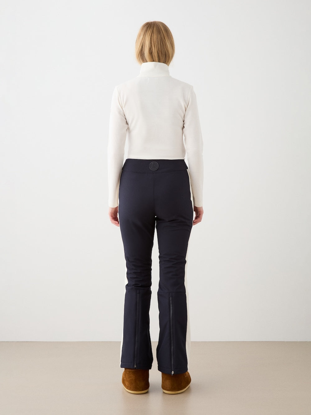 A person with light brown hair, dressed in a white long-sleeve top and We Norwegians Solo Ski Pant women, faces away from the camera against a plain light background.