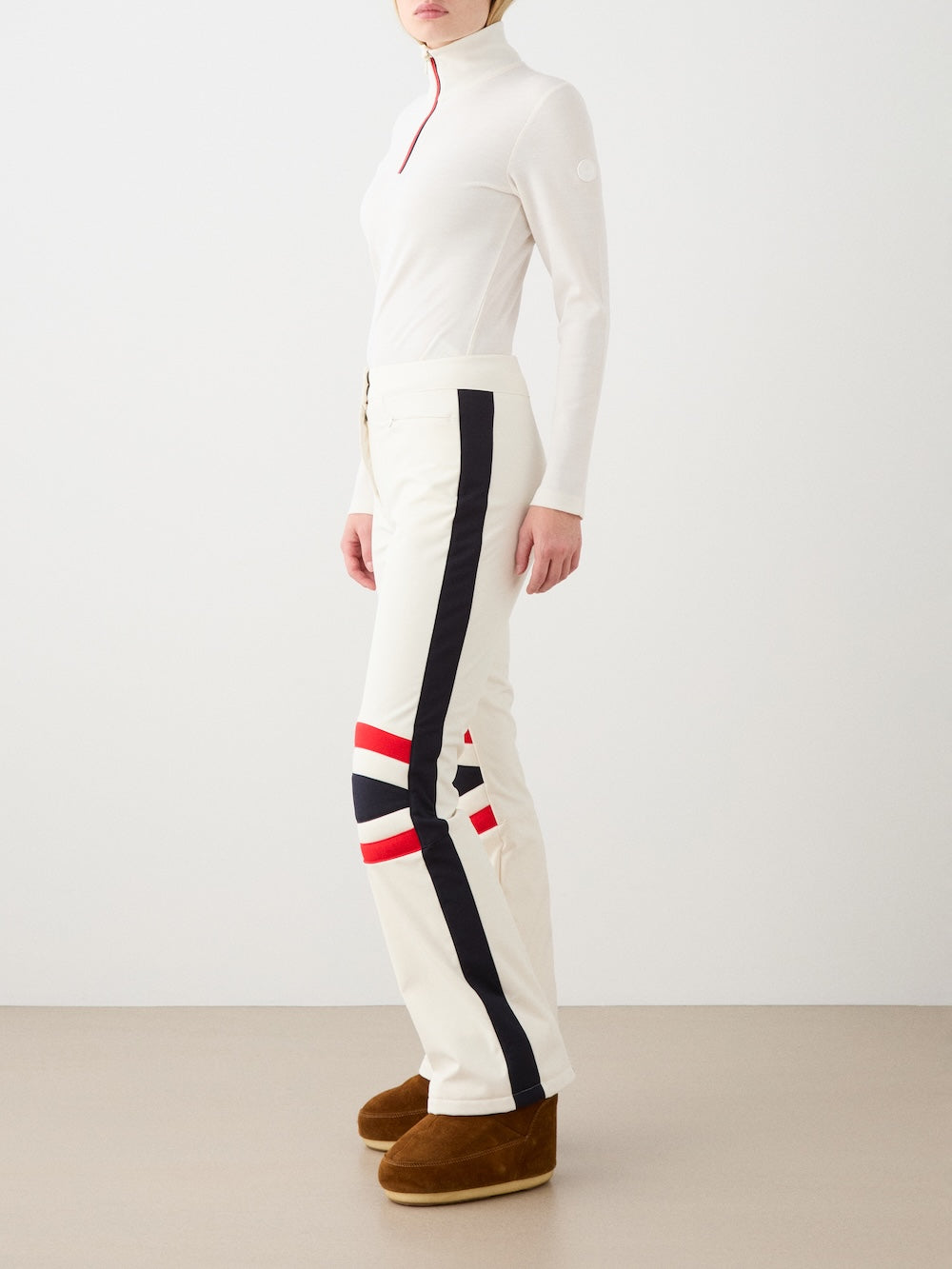 A woman stands sideways against a plain background wearing We Norwegians' Solo Ski Pant, featuring padded knees and black and red side stripes, paired with a fitted white long-sleeve top and brown winter boots.