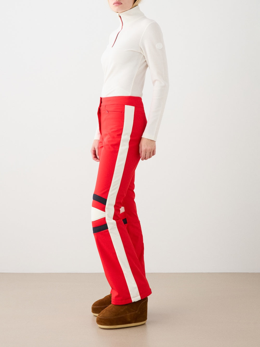 A person wears the We Norwegians Solo Ski Pant for women, featuring white and navy side stripes, paired with a fitted white long-sleeve top and brown ankle boots, standing against a plain background.