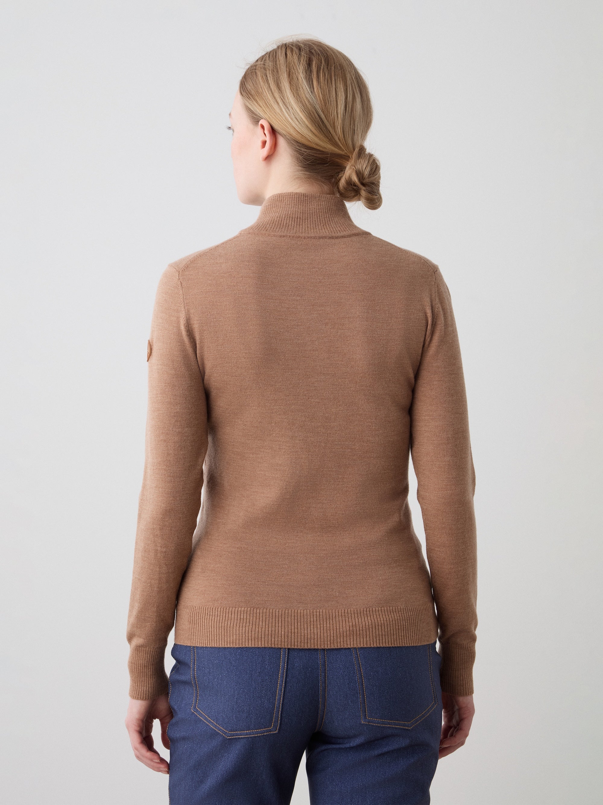 A woman with blonde hair in a low bun wears the We Norwegians FreeBird ZipUp, standing with her back to the camera against a plain light background.