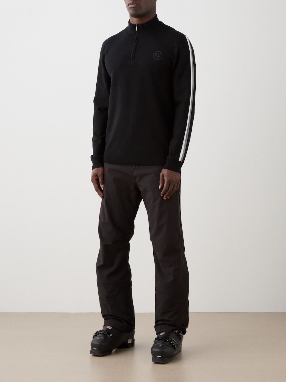 The model is wearing The We Norwegians FreeBird ZipUp men is a black long-sleeve ski sweater with a quarter-zip collar, white stripes on the shoulders and sleeves, and a subtle circular logo on the left chest.