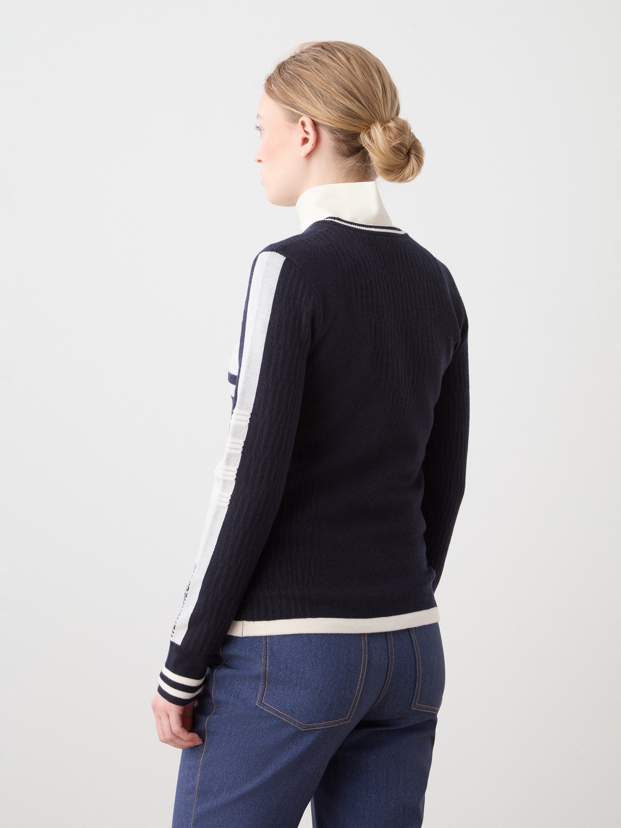 A woman with blonde hair in a low bun is seen from the back, wearing the We Norwegians Rocky Ski Sweater with a white collar and blue jeans, standing against a plain light background.