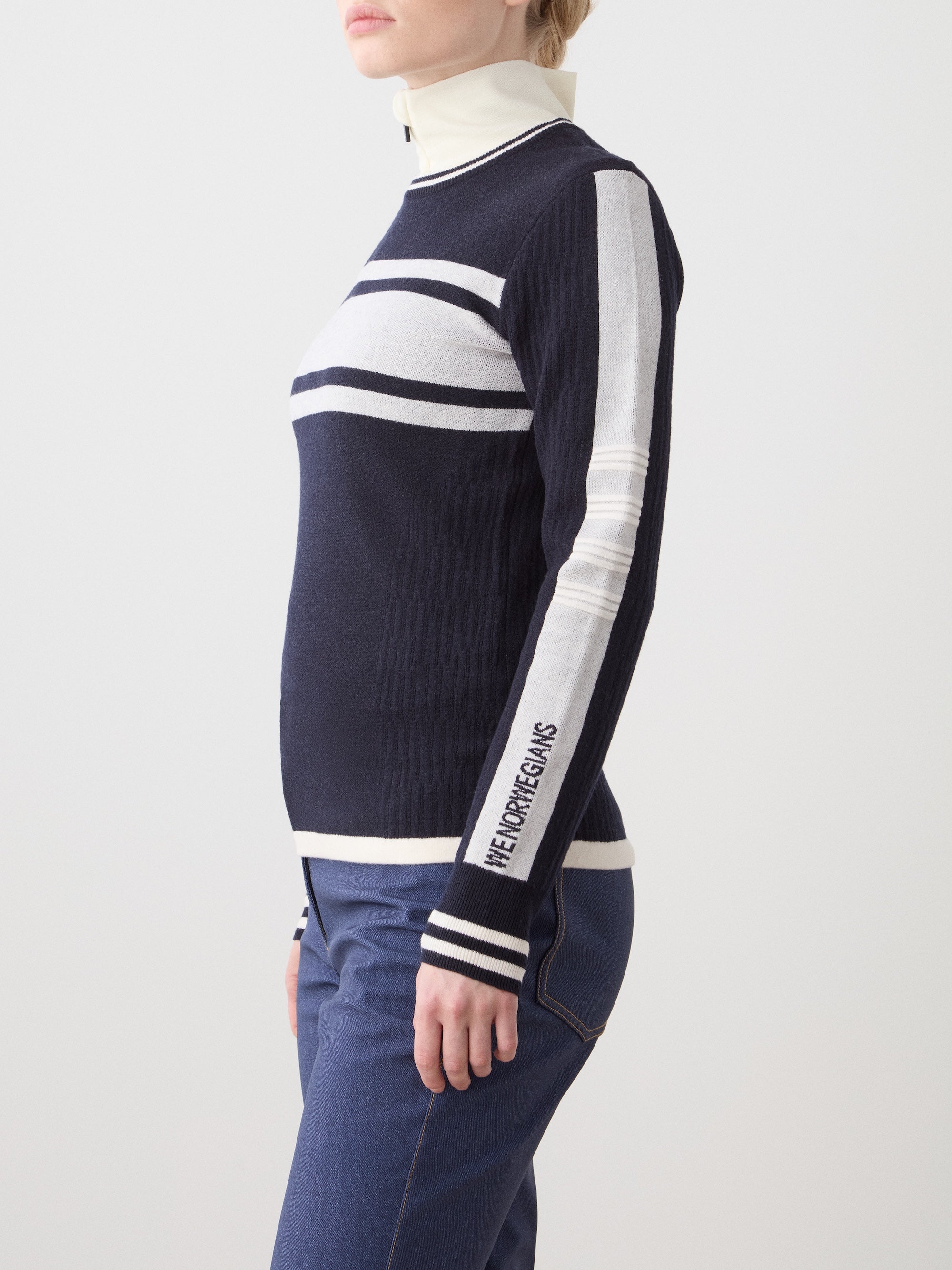 A woman in a navy and white striped We Norwegians Rocky Ski Sweater with "VETEMENTS" on the sleeve, paired with blue jeans, stands against a plain light background.