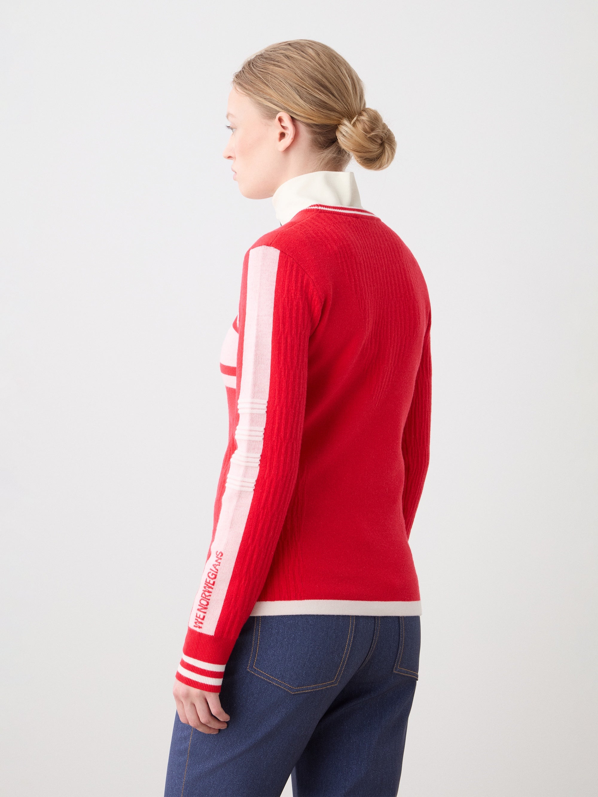 A woman with blonde hair in a low bun wears the We Norwegians Rocky Ski Sweater for women—a red and white retro striped knit—paired with blue jeans, seen from the side and back against a plain light background.