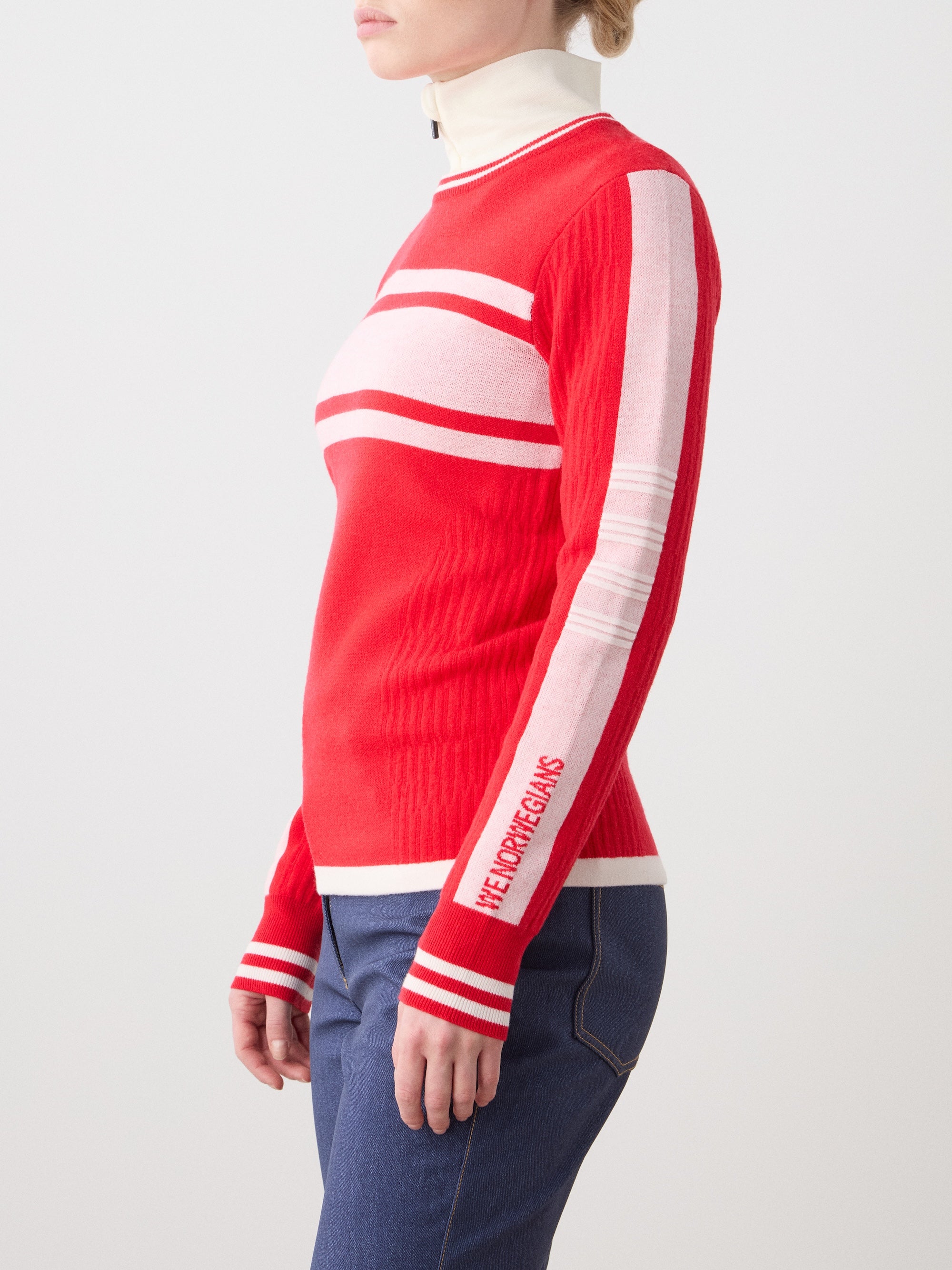A woman in profile wears the We Norwegians Rocky Ski Sweater for women in red and pink with white stripes and "NEW DREAMS" on the sleeve, paired with blue jeans, against a plain background.