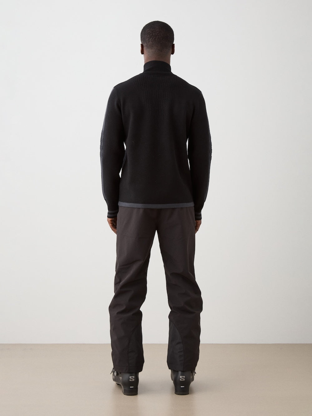 A man stands indoors facing away from the camera, wearing the We Norwegians Rocky FullZip men sweater with dark pants and black shoes, against a plain white wall and a light-colored floor.