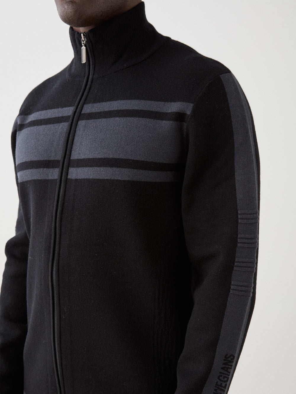 A person wearing the We Norwegians Rocky FullZip men, a black retro racing knit jacket with two dark gray horizontal stripes on the chest and sleeves, high collar, and visible lower face and upper body.