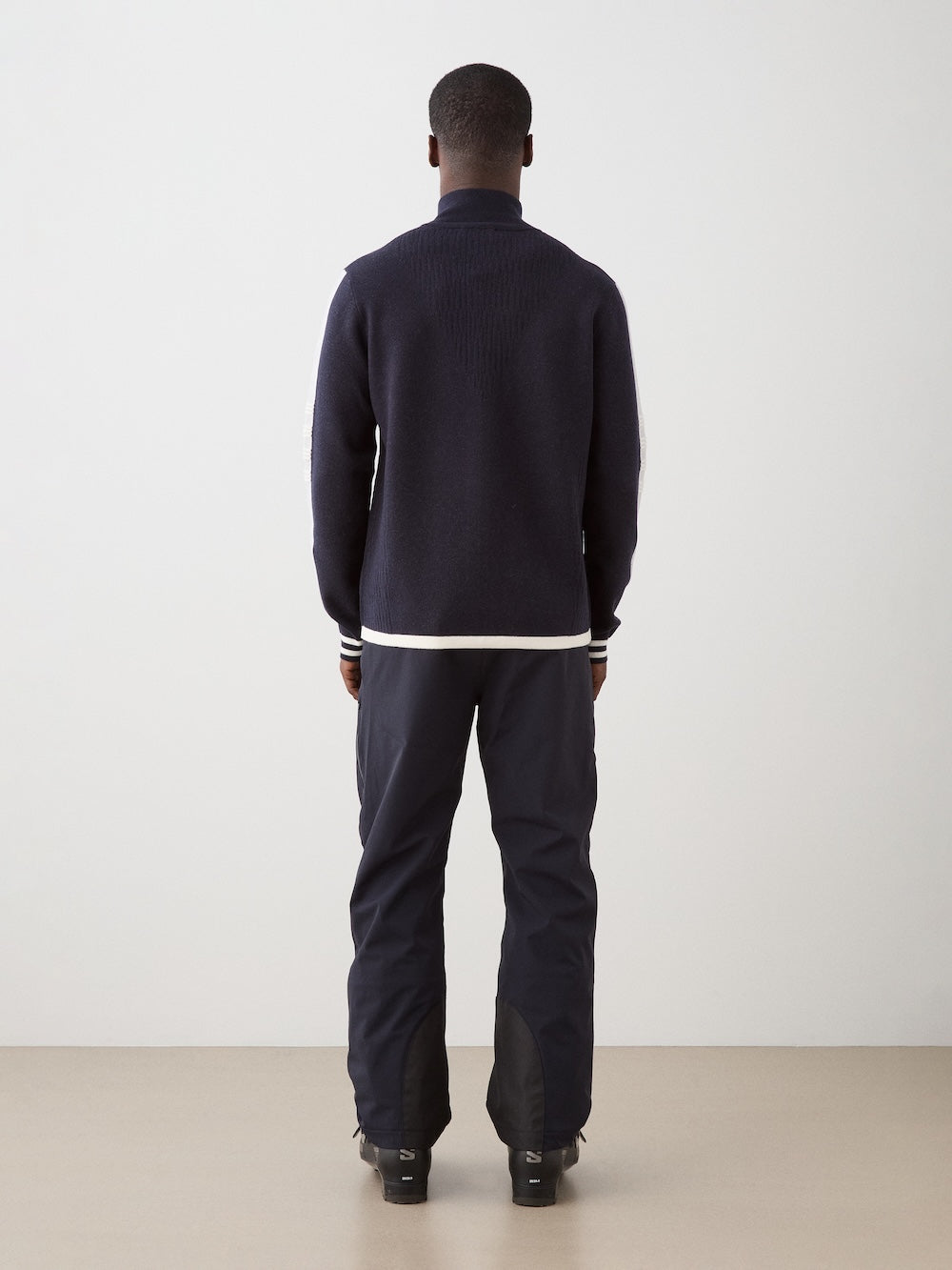 A man, seen from behind, wears the We Norwegians Rocky FullZip with dark pants in a minimal indoor space featuring a plain light-colored wall and floor.