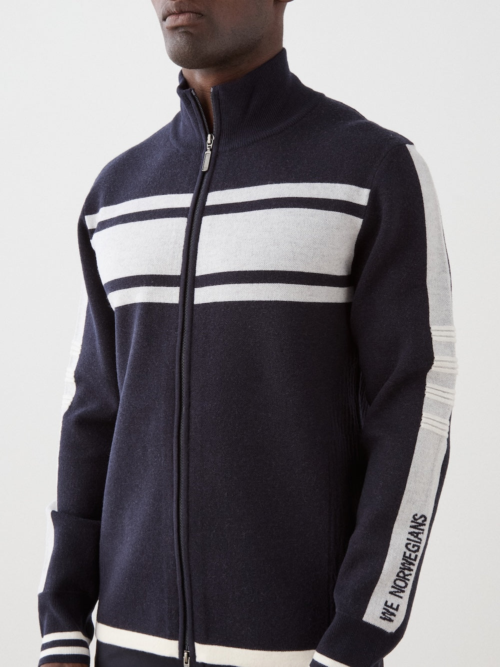 A man wears the We Norwegians Rocky FullZip jacket in navy blue merino wool, featuring white stripes, "WE NORWEGIANS" text on the left sleeve, a high collar, and a modern retro racing knit design.