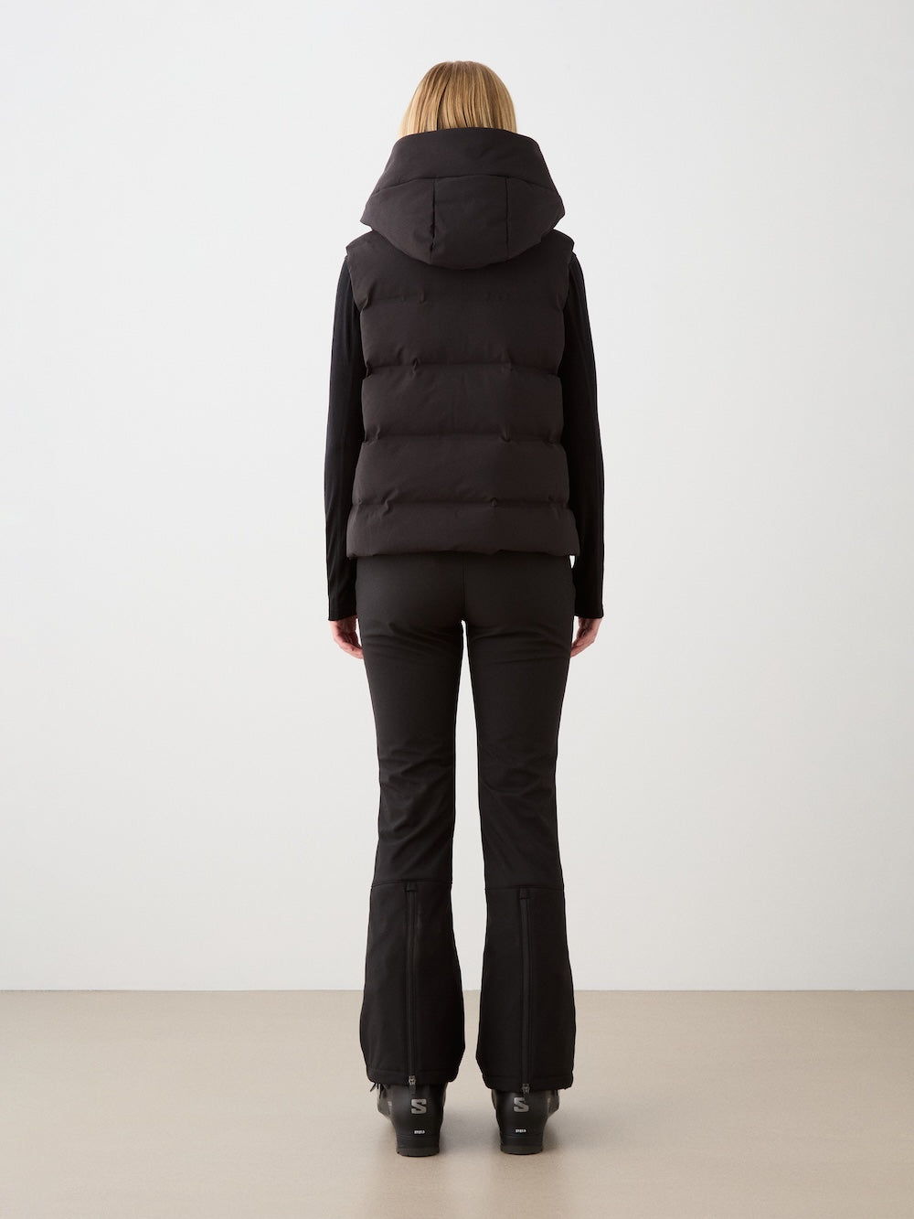 A person faces away, wearing the We Norwegians Aurora Vest for women—a black hooded responsible down vest—styled with black fitted pants and shoes, capturing a minimalist Scandinavian look against a plain light backdrop.