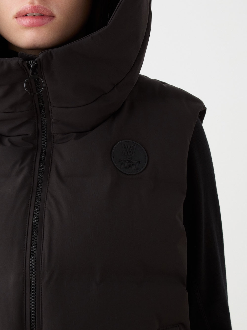 A woman wears the We Norwegians Aurora Vest—a black hooded puffer with a zipper and circular logo—capturing Scandinavian style, shown from the shoulders up against a plain light background.