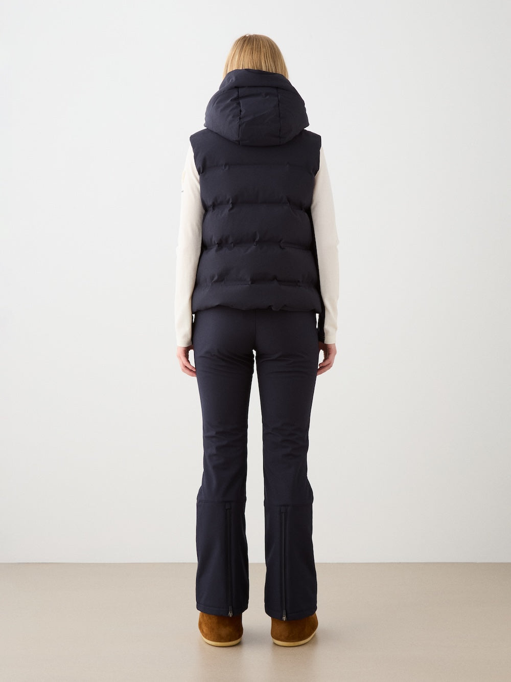 A person faces away, wearing We Norwegians’ Aurora Vest women—a black, responsibly sourced down vest with a hood—styled with a light long-sleeve top, black fitted pants, and brown shoes. The plain background evokes Scandinavian minimalism.