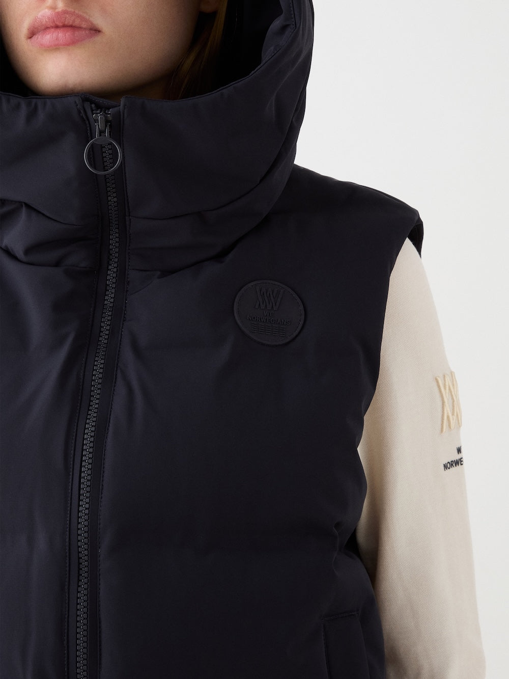 A close-up of a person wearing the We Norwegians Aurora Vest women—a black, sleeveless puffer with high collar and zipper—over a light long-sleeve shirt with embroidered logos, reflecting a refined Scandinavian style.