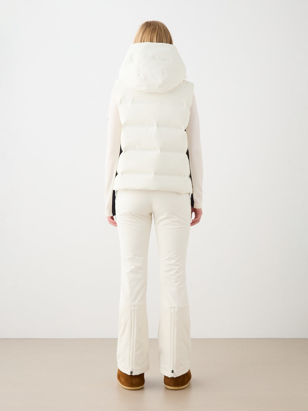 A woman faces away, wearing the We Norwegians Aurora Vest women—a responsible down hooded vest—in all white with fitted pants and brown boots, creating a clean Scandinavian look against a plain light background.