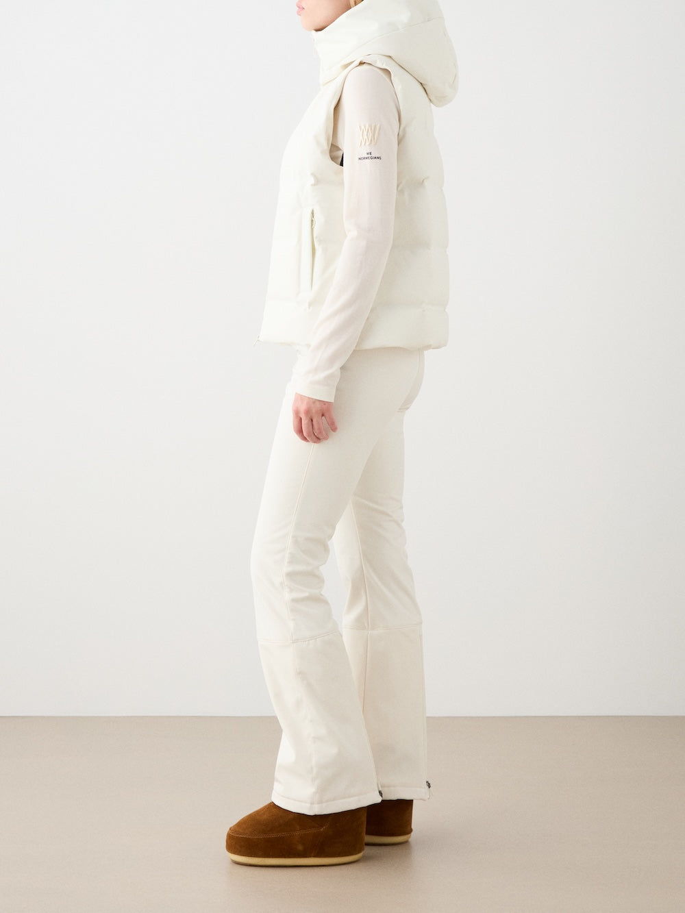 A woman stands sideways against a plain background in an all-white look, showcasing the We Norwegians Aurora Vest women—a responsible down vest with a clean, Scandinavian style—paired with a white shirt, flared pants, and brown winter boots.