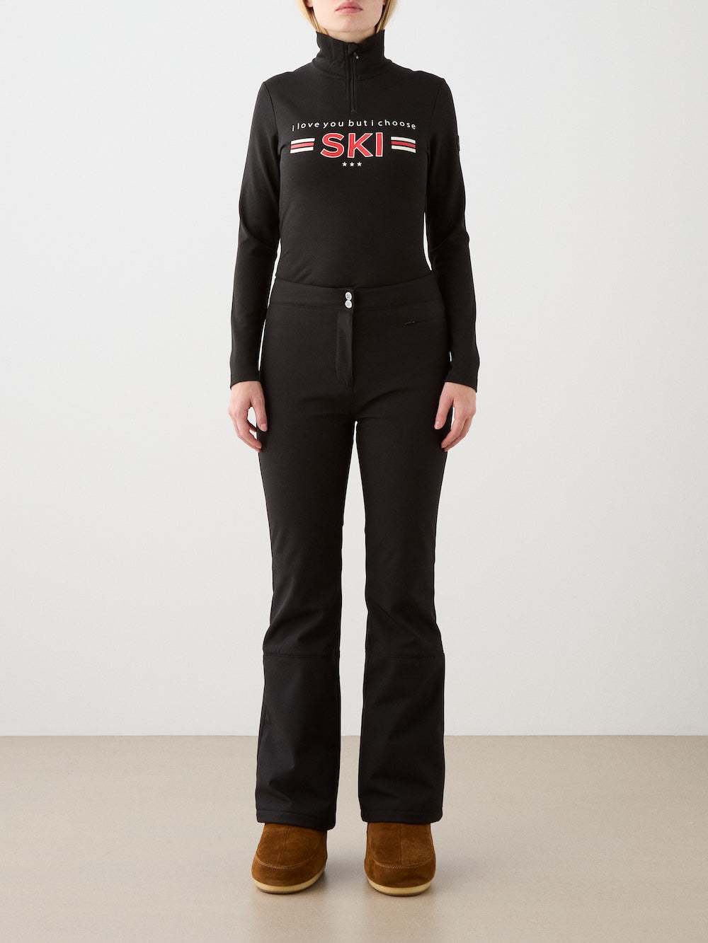 A person stands facing forward in the We Norwegians Bibi ZipUp women—a black merino wool top with “I love you but I choose SKI”—paired with black fitted pants and brown slip-on boots against a plain light background, evoking vintage ski style.