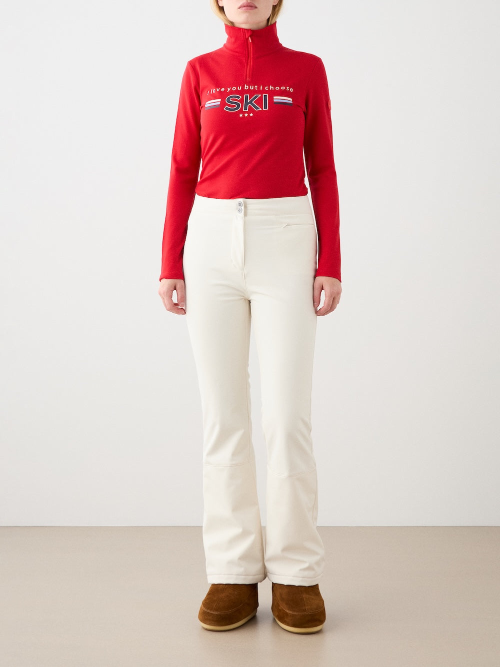 A person stands against a neutral background, wearing the We Norwegians Bibi ZipUp women red merino wool top featuring "I like you but I choose SKI," paired with off-white pants and brown winter boots. Their hands rest by their sides.