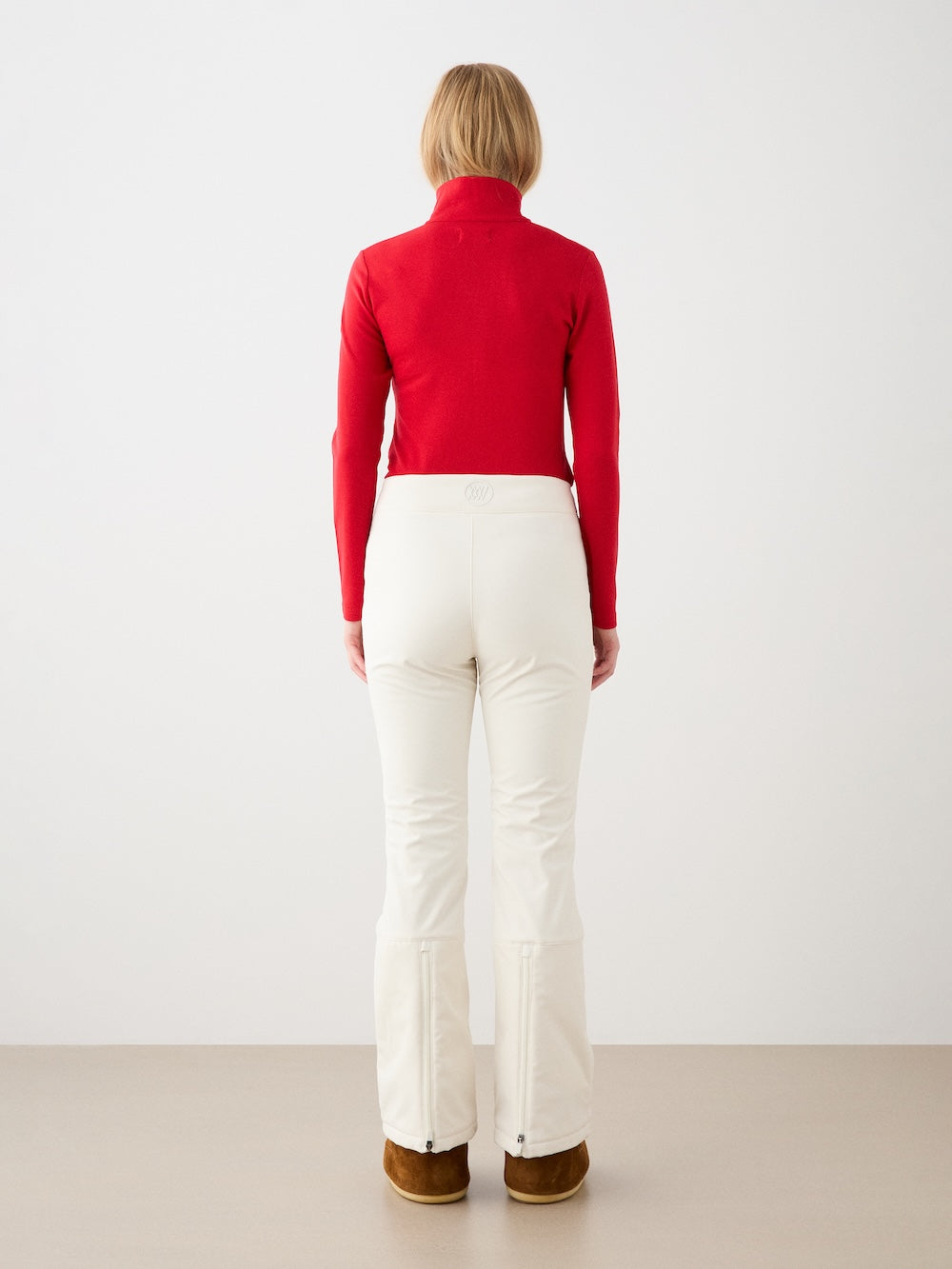 A person with short blond hair models the We Norwegians Bibi ZipUp women, wearing a red merino wool top, white pants, and brown shoes, standing facing away from the camera against a plain light background.