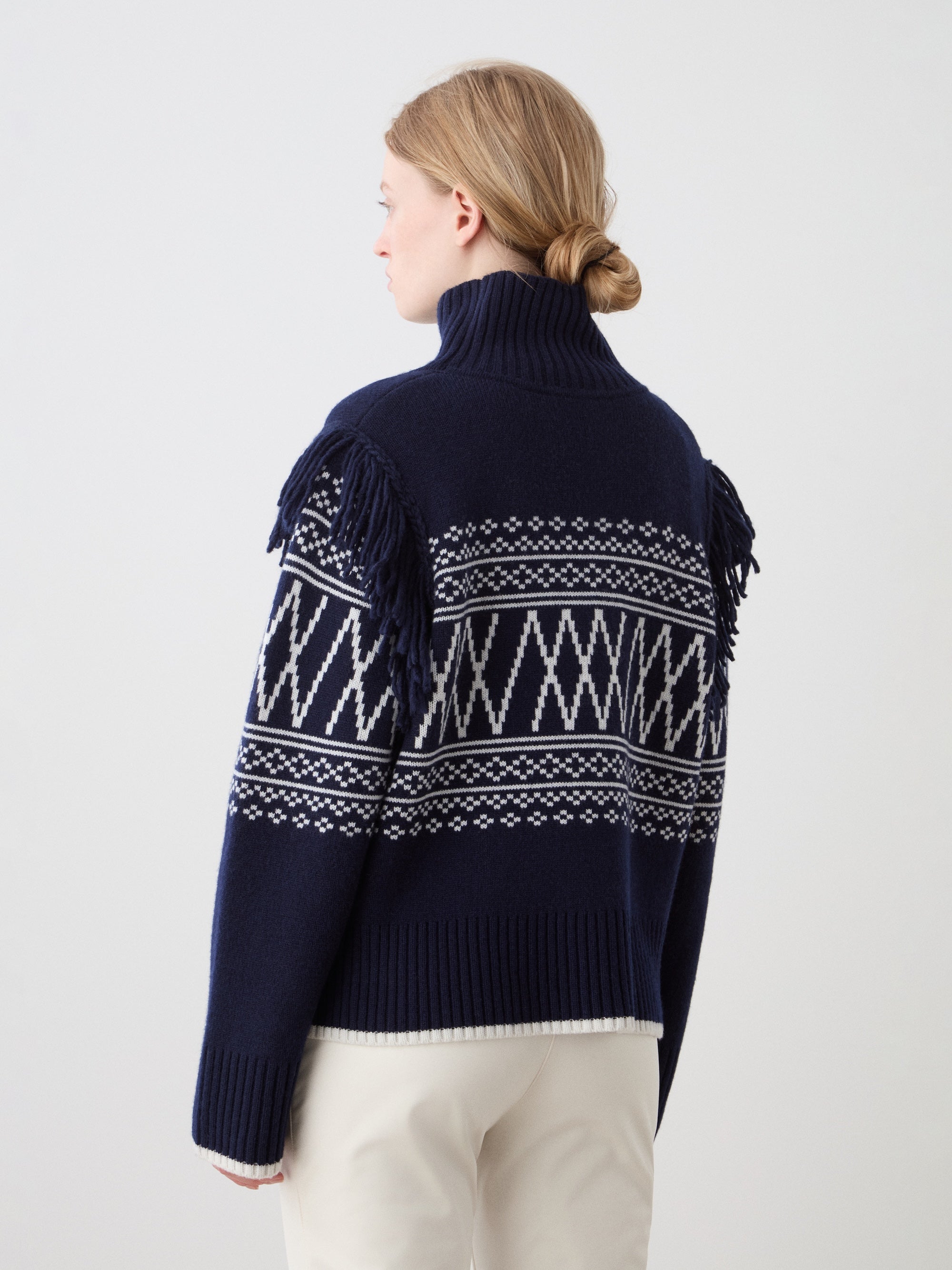 A woman with light hair in a low bun wears the We Norwegians Setesdal Sweater women in navy with white geometric patterns and shoulder fringe, paired with light pants, posing against a plain background.