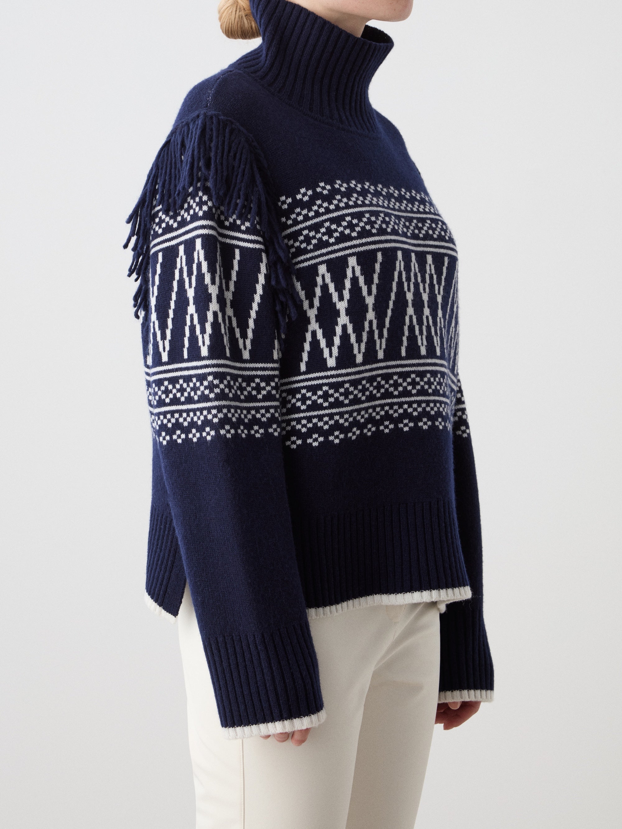 A woman wears the We Norwegians Setesdal Sweater in navy blue with white geometric patterns and fringe shoulders, made from a soft merino-cashmere blend, paired with off-white pants against a plain light background.