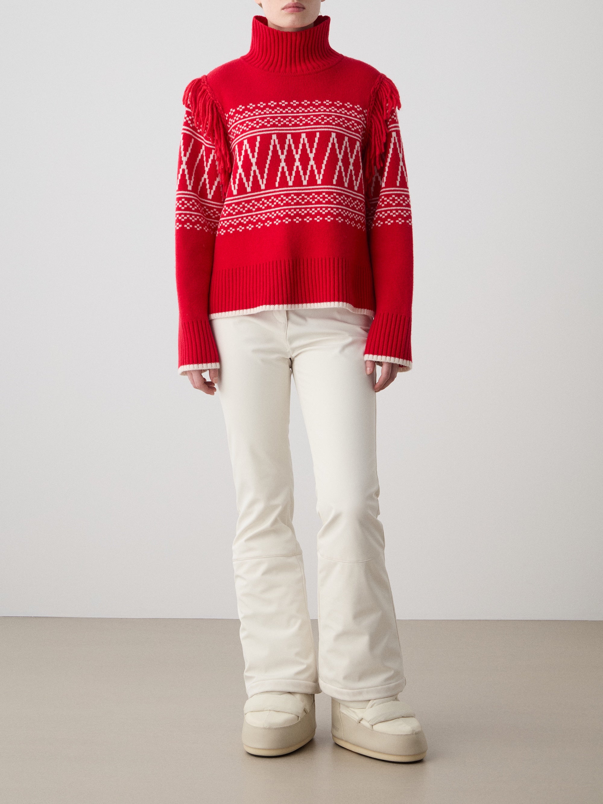 A woman stands indoors against a plain background wearing the We Norwegians Setesdal Sweater, a red Norwegian knit turtleneck with white geometric patterns, paired with cream-colored flared pants and chunky off-white boots.