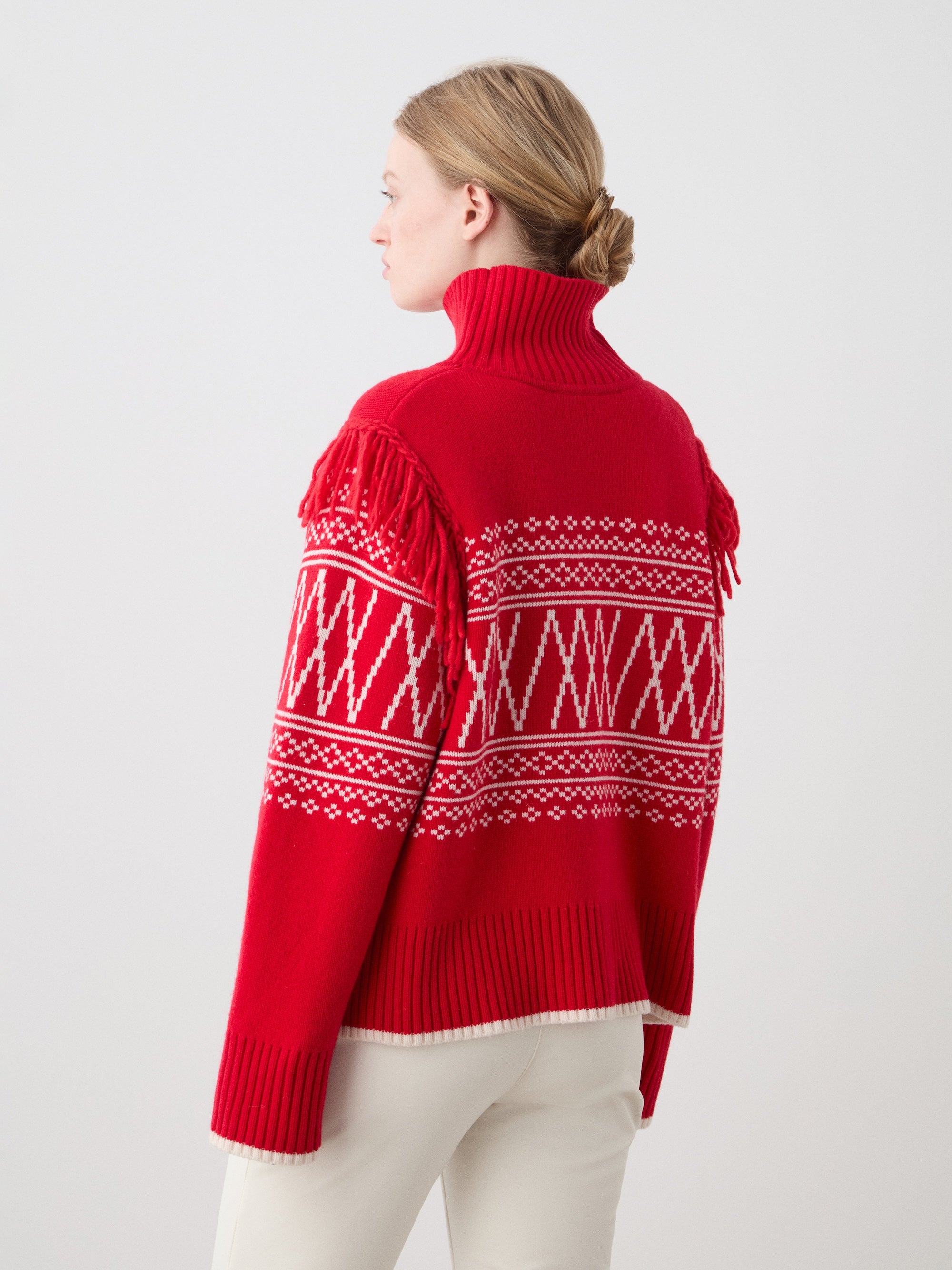 A woman with light skin and blonde hair in a low bun faces away from the camera, wearing the We Norwegians Setesdal Sweater for women in red with white geometric patterns and shoulder fringes, paired with light-colored pants.