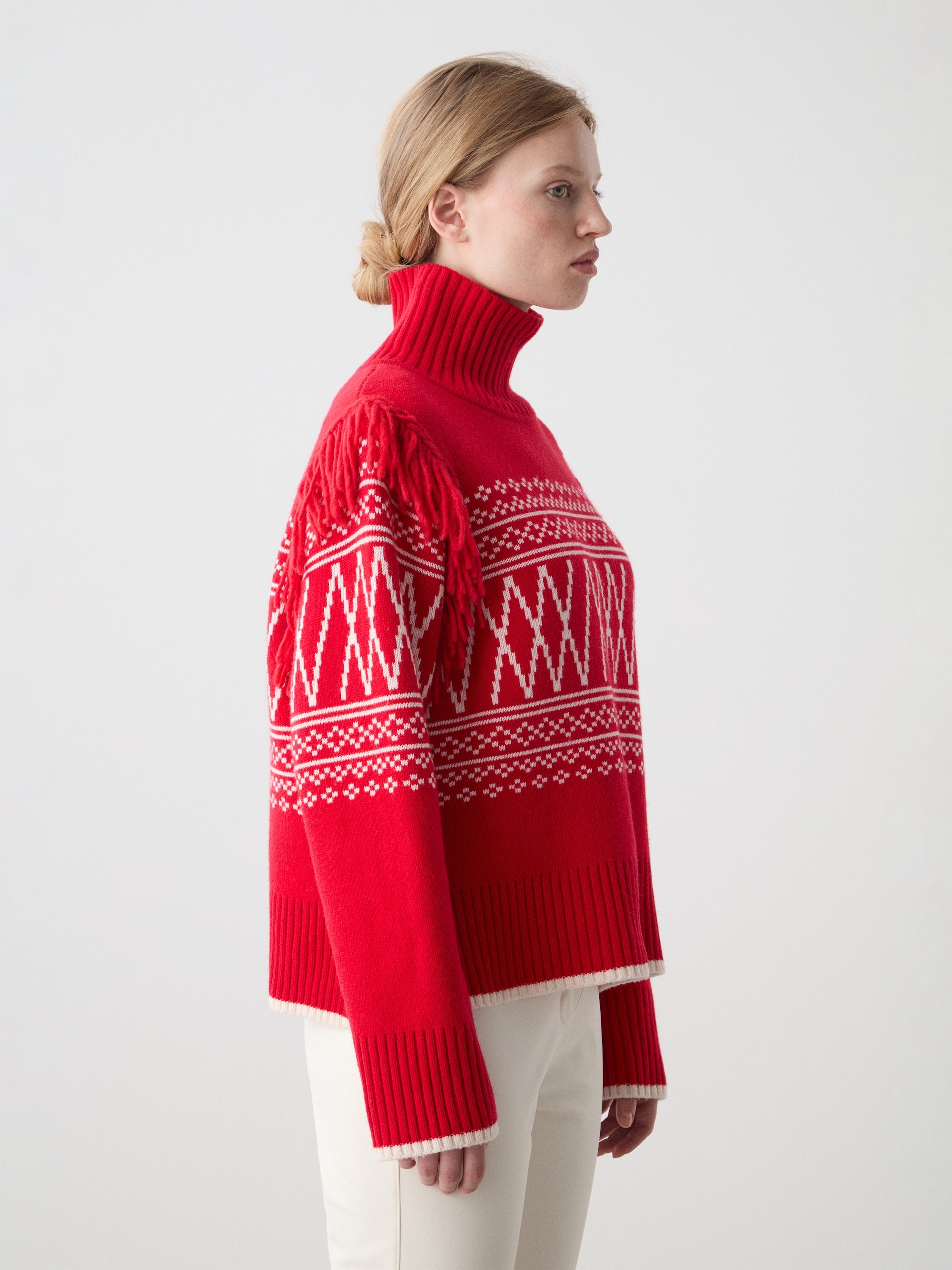A woman with blonde hair in a low bun models the We Norwegians Setesdal Sweater for women, a red turtleneck with white geometric patterns, paired with cream pants and set against a plain light background.