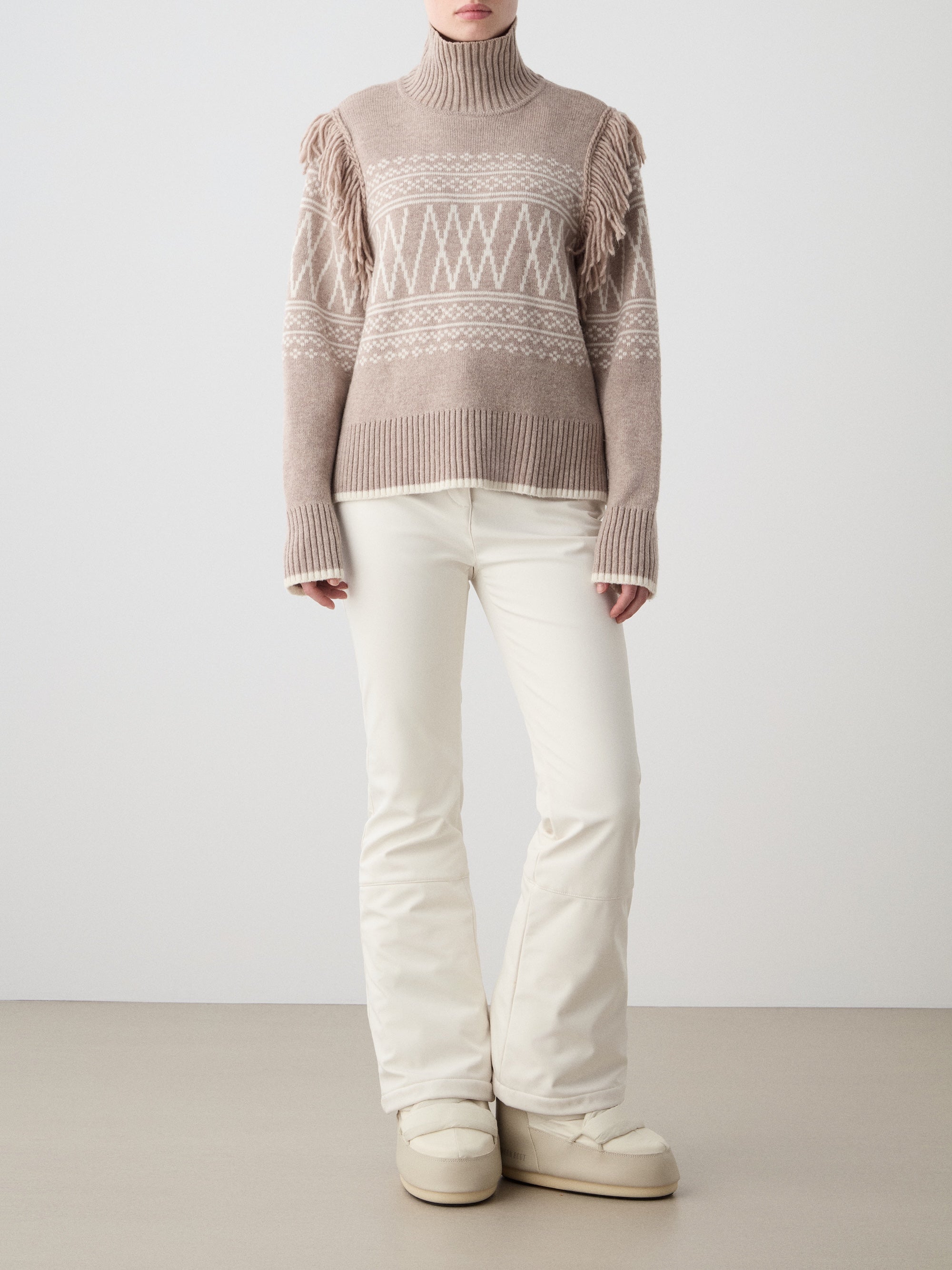 A person wears the We Norwegians Setesdal Sweater women—a beige knit with white geometric patterns and fringed shoulders—paired with off-white pants and beige boots, standing against a plain light background.
