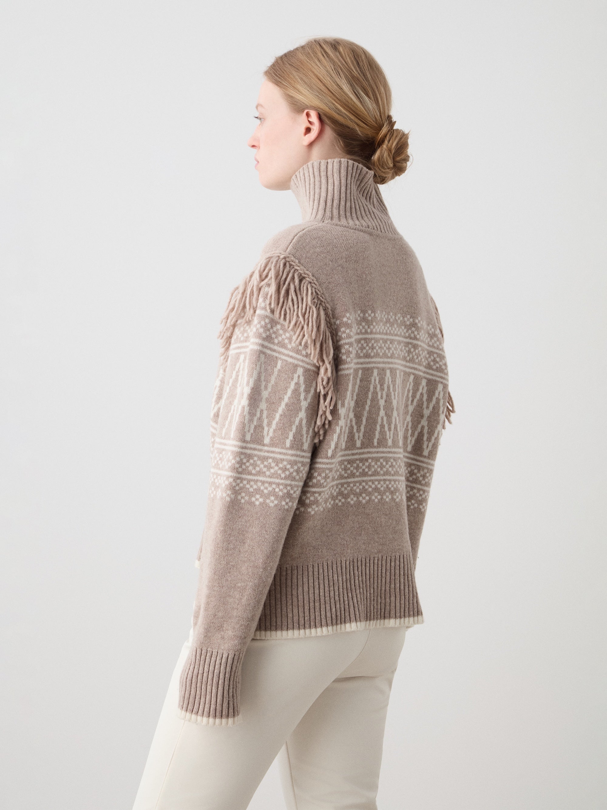 A woman with blonde hair in a low bun wears the We Norwegians Setesdal Sweater for women—a beige and white patterned merino-cashmere blend with shoulder fringe—paired with cream pants against a plain light background.