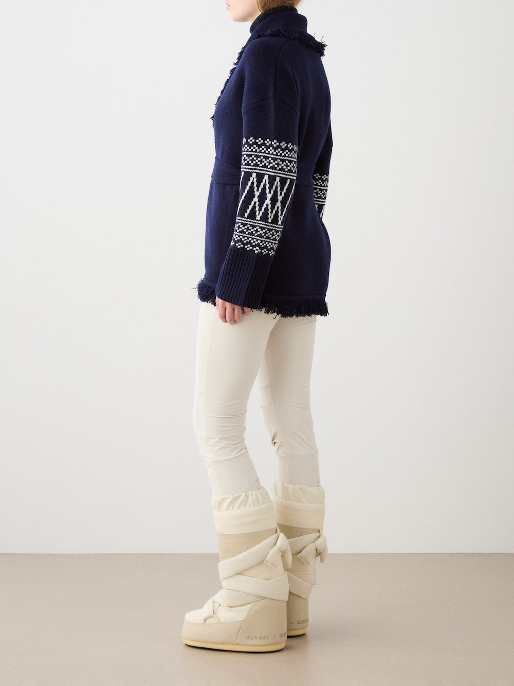 A woman stands sideways against a plain background wearing the We Norwegians Setesdal Cardigan in navy blue with white patterned sleeves, plus white fitted pants and beige snow boots. Her face is partially out of the frame.