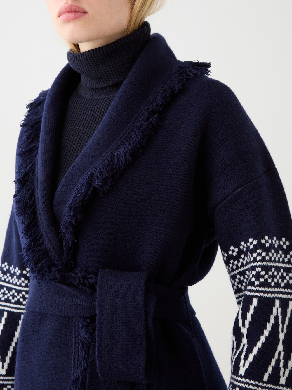 A person wearing the We Norwegians Setesdal Cardigan women in navy blue with a fringed collar, white Norwegian patterns on the sleeves, a matching belt, and a black ribbed turtleneck underneath. Photo cropped at mouth and chest.