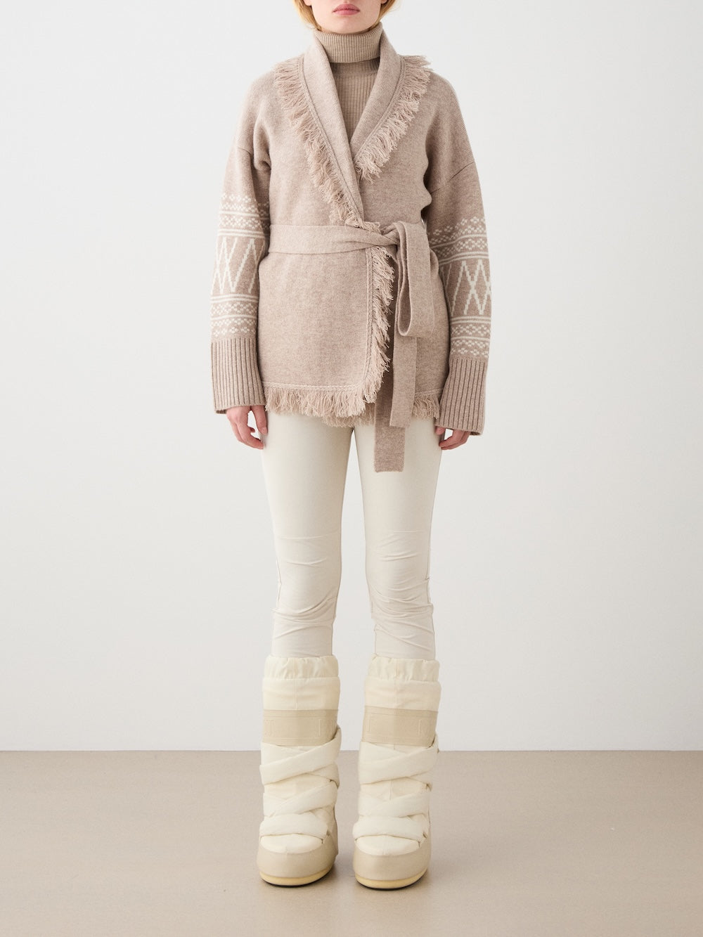A woman wearing the We Norwegians Setesdal Cardigan—a beige belted cardigan featuring fringe and patterned sleeves inspired by classic Norwegian knits—paired with a tan turtleneck, white pants, and cream-colored snow boots, stands against a plain light background.