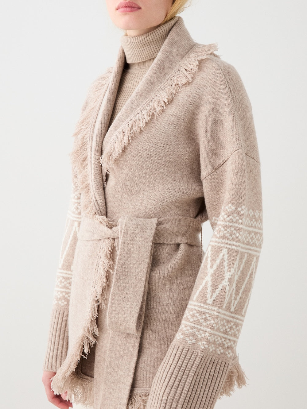 A person wearing the We Norwegians Setesdal Cardigan women, a beige belted merino-cashmere piece with fringed edges and white geometric sleeve patterns, over a matching turtleneck, stands against a plain white background.