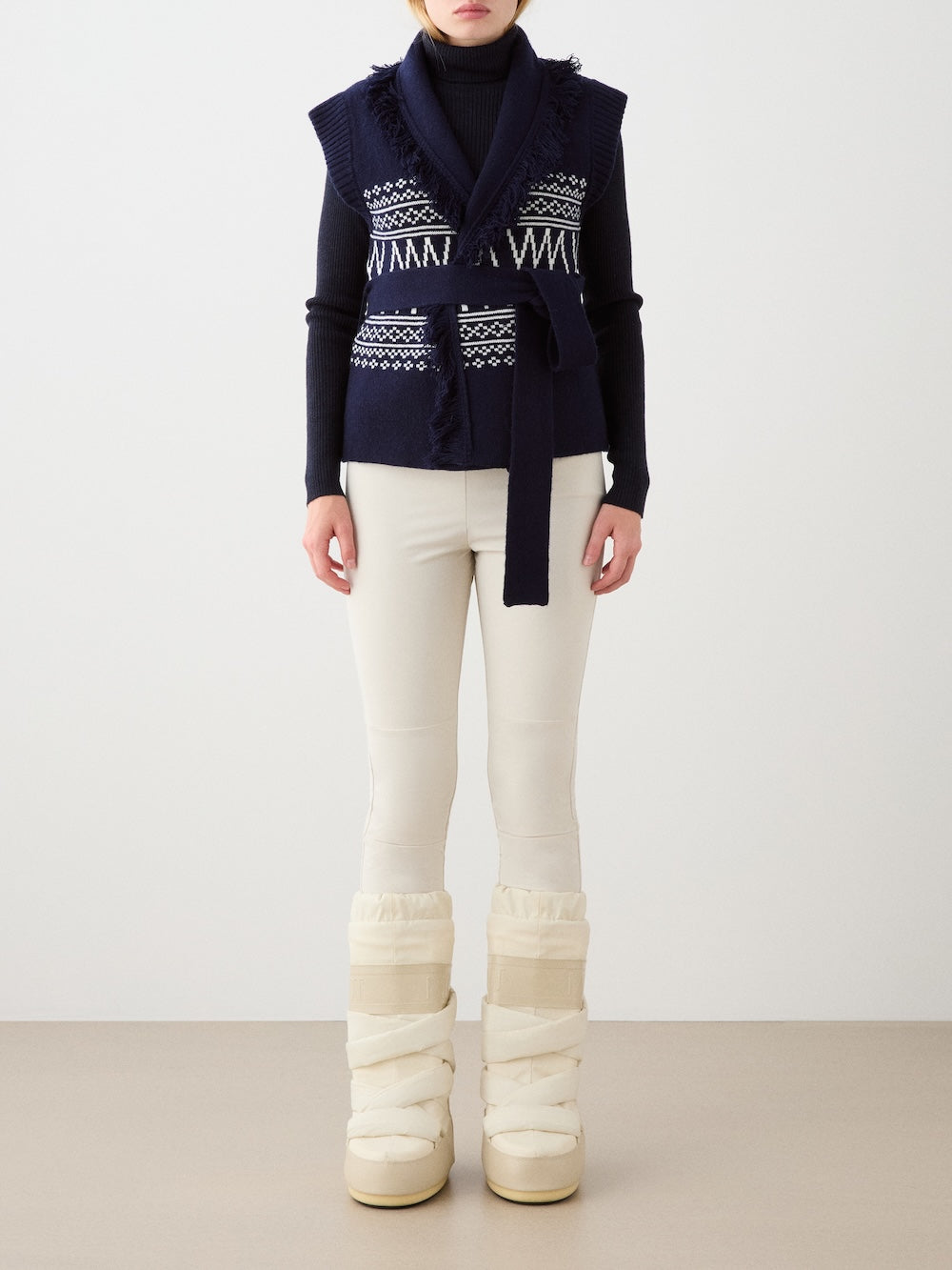 A person models the We Norwegians Setesdal Vest women in navy blue over a black turtleneck, paired with cream fitted pants and tall winter boots, standing against a plain background. The face is not visible.