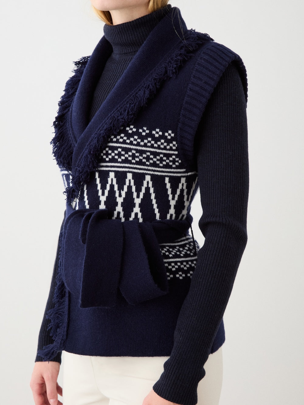 A side view of a woman in the We Norwegians Setesdal Vest—navy blue with white geometric patterns, fringed shawl collar, and matching belt—layered over a ribbed navy turtleneck and white pants for a classic après-ski look.