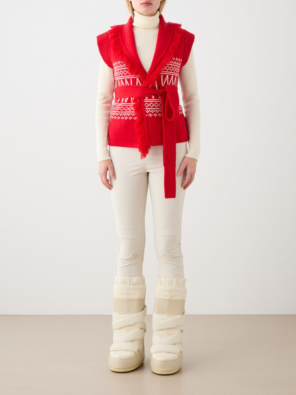 A model wears the We Norwegians Setesdal Vest for women—red patterned, sleeveless, merino-cashmere with a belt—over a white turtleneck sweater, cream pants, and tall cream winter boots, showcasing chic après-ski style against a white background.