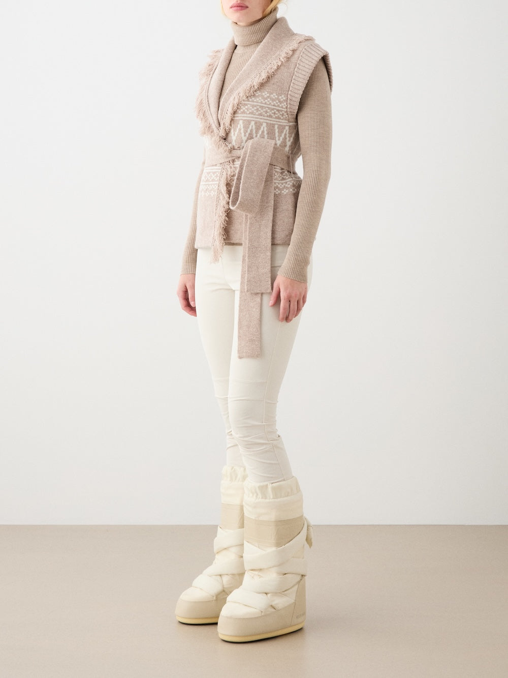A person wearing the We Norwegians Setesdal Vest women—a beige merino-cashmere vest with textured details—over a beige turtleneck, white pants, and cream winter boots stands against a plain background. Their face is not visible.