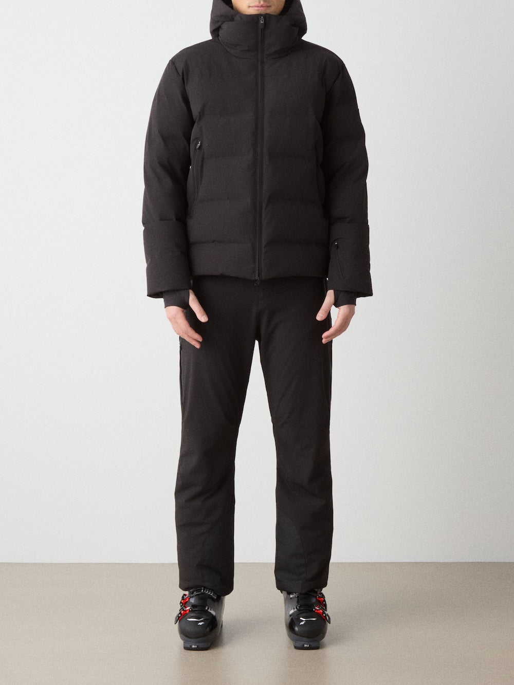 A person stands facing forward in a We Norwegians Nordlys Ski Jacket men—black, waterproof, hooded, with responsible down—paired with black pants and black ski boots with red accents. Face partially out of frame; background is plain.